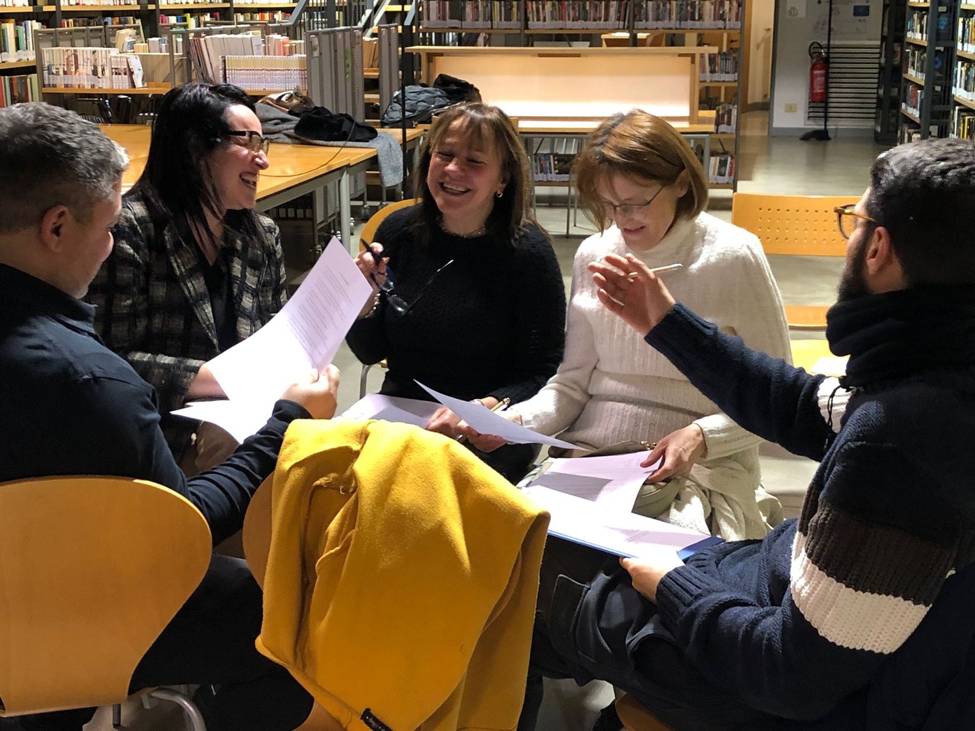 Un gruppo di partecipanti ride confrontandosi sulle schede, attorno a un tavolo della biblioteca